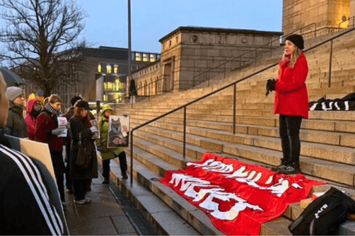 Punatakkinen Salla eduskuntatalon portailla edessään joukko ihmisiä mielenosoituskylttien kanssa.