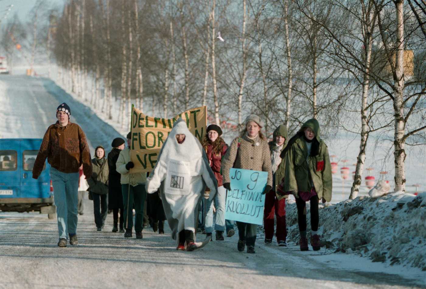 Pieni mielenosoituskulkue talvisessa maisemassa, eturivissä Salla Tuomivaara kädessään kyltti, jossa lukee "jo 9 delfiiniä kuollut".