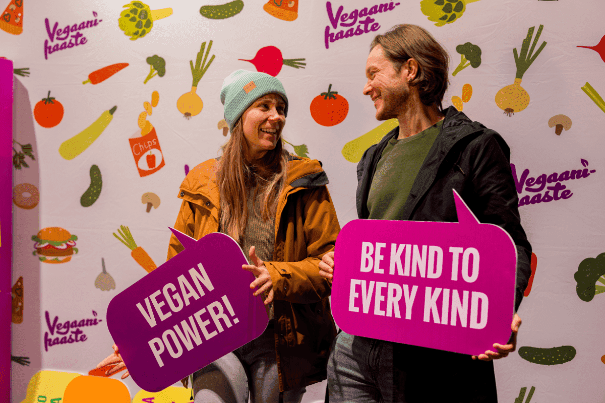 Kaksi hymyilevää ihmistä katsoo toisiinsa kuvausseinän edessä käsissään kyltit, joissa lukee "Vegan power!" ja "Be kind to every kind"